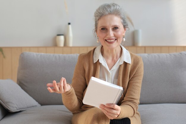 Jolie femme mûre assise sur le canapé et prenant des notes dans le journal.