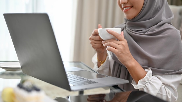 Jolie femme musulmane asiatique recadrée en sirotant un café du matin tout en utilisant un ordinateur portable