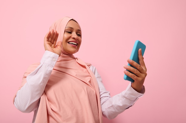 Jolie femme musulmane d'ethnie moyen-orientale ou arabe avec la tête couverte en hijab rose ayant une vidéoconférence sur téléphone portable, saluant son interlocuteur lors d'un appel vidéo, espace de copie sur fond rose