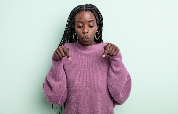 Jolie femme noire avec la bouche ouverte pointant vers le bas avec les deux mains, l'air choquée, étonnée et surprise