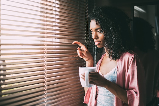 Jolie femme noire debout à la fenêtre avec une tasse de café