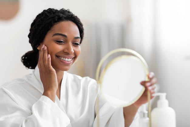 Jolie femme noire regardant un miroir et touchant une peau parfaite sur le visage