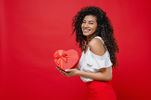 Jolie femme noire souriante tenant cadeau en forme de coeur isolé sur rouge