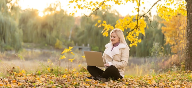 Jolie femme avec ordinateur portable dans le parc en automne. Scène de nature de beauté avec fond de feuillage coloré, arbres jaunes et feuilles à l'automne. Mode de vie en plein air d'automne. Heureuse femme souriante sur les feuilles d'automne