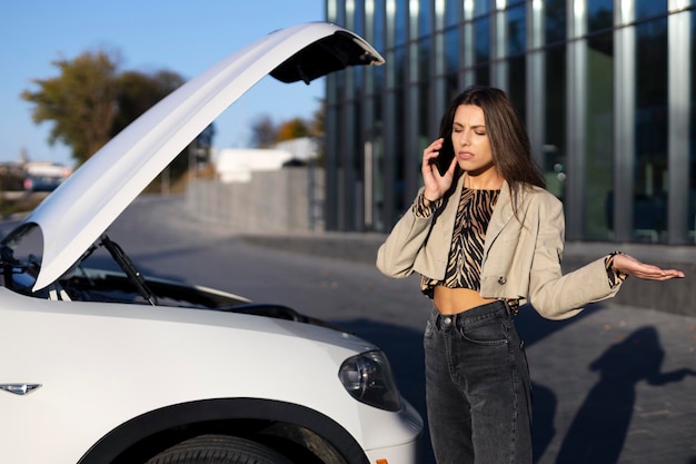 Jolie femme parlant au téléphone debout près d'une voiture avec un capot ouvert écartant ses mains à l'extérieur