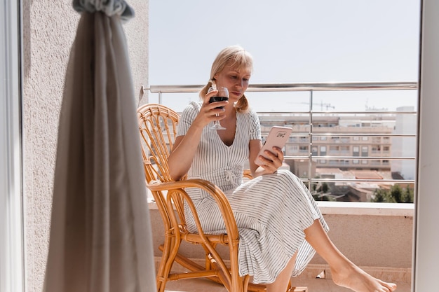 Une jolie femme parle au téléphone sur la terrasse ensoleillée avec un verre de vin à la main