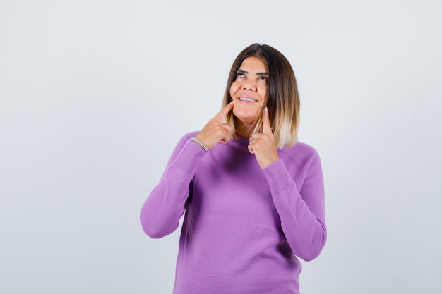 Jolie femme pointant sur ses fossettes, levant les yeux dans un pull violet et l'air gaie, vue de face.