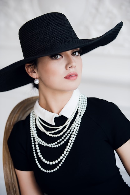 Jolie femme portant une robe noire, un chapeau et des perles, assis sur une chaise