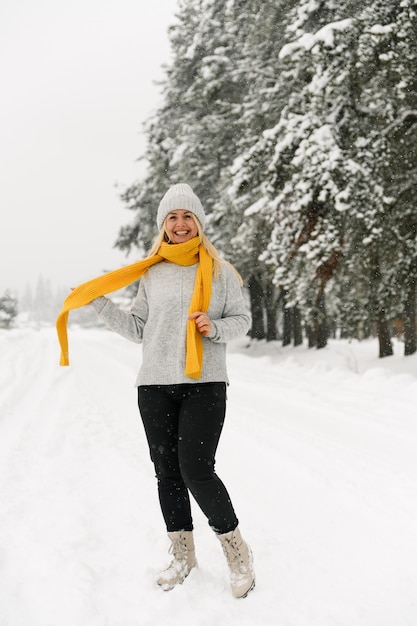 Jolie femme en pull gris et écharpe jaune marche dans une chute de neige. Femme s'amusant dans la forêt d'hiver. Portrait en plein air de la beauté blonde lady posing in wood