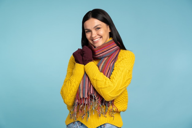 Jolie femme en pull jaune élégant sourit largement. Fille cool en écharpe tricotée chaude et gants posant sur fond bleu. Concept d'hiver