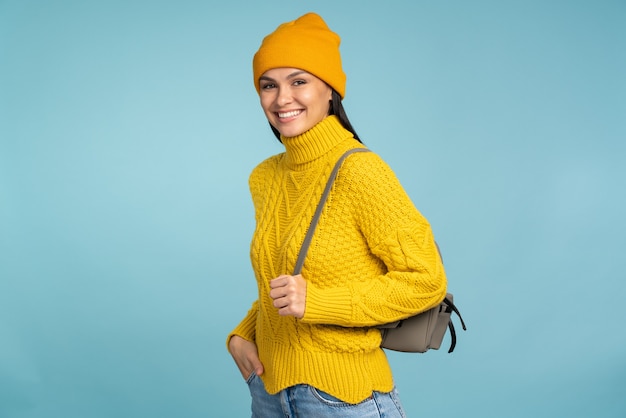 Jolie femme en pull jaune élégant sourit largement en posant avec son sac à dos. Fille cool dans des vêtements tricotés chauds, debout sur le fond bleu. Concept d'hiver