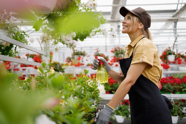 Jolie femme pulvérisant des fleurs avec un flacon pulvérisateur en serre