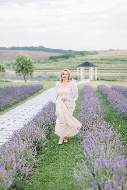 Jolie femme de race blanche blonde mature avec une robe beige clair qui traverse joyeusement un champ de lavande souriant à la caméra