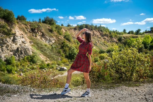 Jolie femme en robe à carreaux rouge chic posant dans la nature, journée ensoleillée d'été