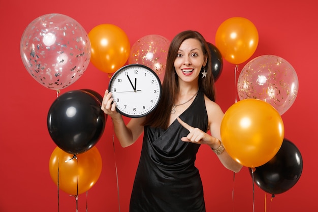 Jolie femme en robe noire célébrant l'index pointé sur une horloge ronde dans les mains sur des ballons à air de fond rouge vif. Saint-Valentin, bonne année, concept de fête de vacances de maquette d'anniversaire.