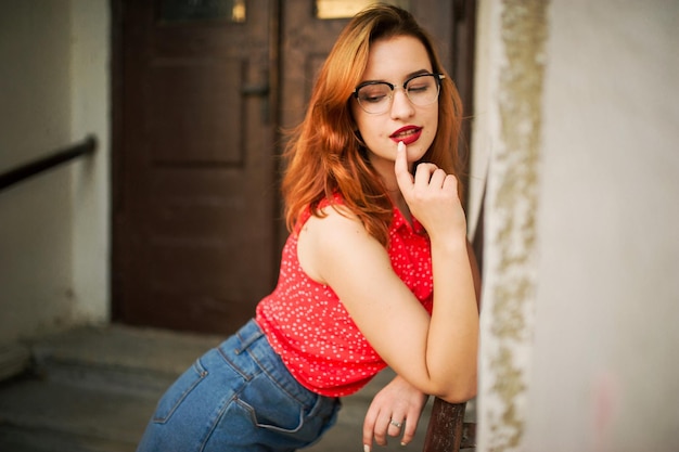 Jolie femme rousse à lunettes porter sur un chemisier rouge et une jupe en jean posant