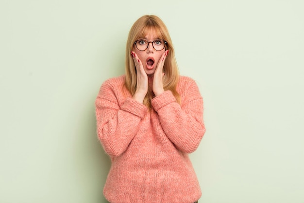 Jolie femme rousse se sentant choquée et effrayée, terrifiée avec la bouche ouverte et les mains sur les joues