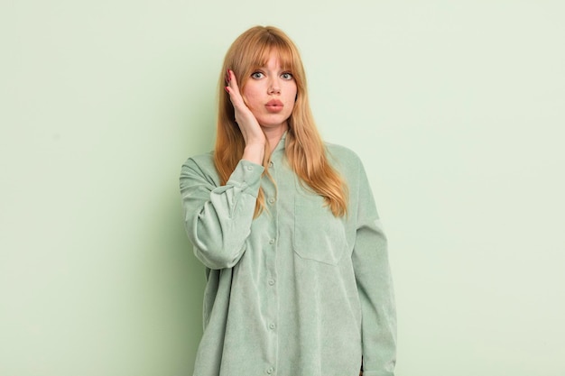 Jolie femme rousse se sentant choquée et étonnée tenant face à main dans l'incrédulité avec la bouche grande ouverte