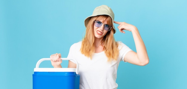 jolie femme rousse se sentant confuse et perplexe, montrant que vous êtes fou et tenant un réfrigérateur portable. concept d'été