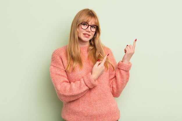 Jolie femme rousse souriant joyeusement et pointant vers le côté et vers le haut avec les deux mains montrant l'objet dans l'espace de copie