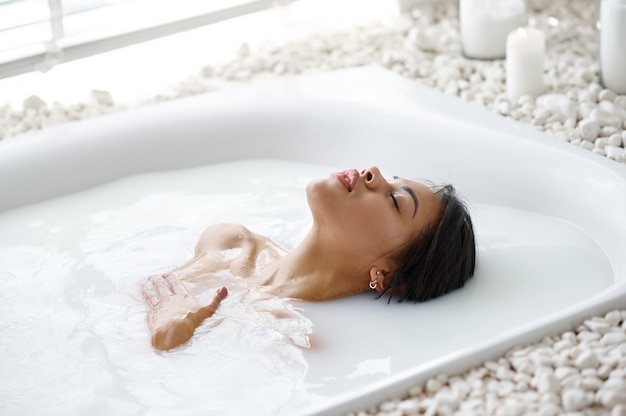 Jolie femme se détendre, soins de la peau dans le bain avec du lait. Personne de sexe féminin dans la baignoire, soins de beauté et de santé au spa, traitement de bien-être dans la salle de bain, cailloux et bougies sur fond