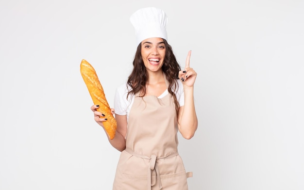 Jolie femme se sentant comme un génie heureux et excité après avoir réalisé une idée portant un tablier et tenant une baguette de pain