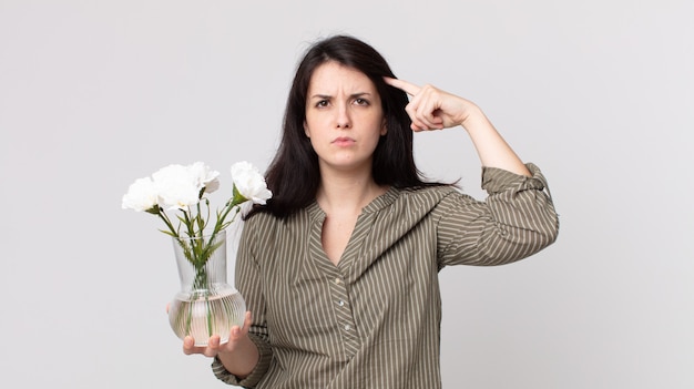 Jolie femme se sentant confuse et perplexe, vous montrant que vous êtes fou et tenant des fleurs décoratives. agent assistant avec un casque