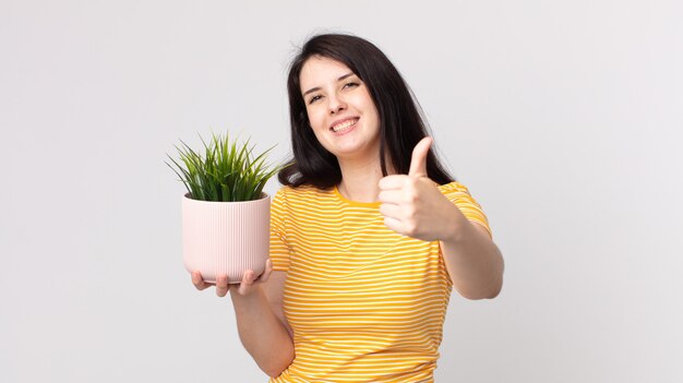 Jolie femme se sentant fière, souriante positivement avec le pouce levé et tenant une plante décorative