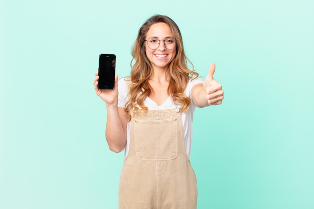 Jolie femme se sentant fière, souriante positivement avec le pouce levé et tenant un smartphone