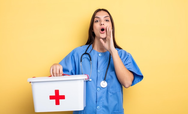 Jolie femme se sentant heureuse de donner un grand cri avec les mains à côté de la bouche infirmière avec une trousse de premiers soins infirmière