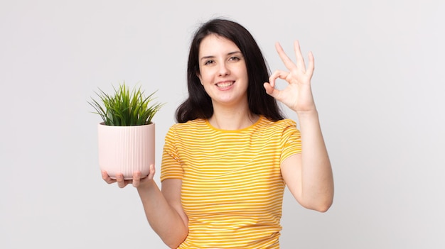 Jolie femme se sentant heureuse, montrant son approbation avec un geste correct et tenant une plante décorative