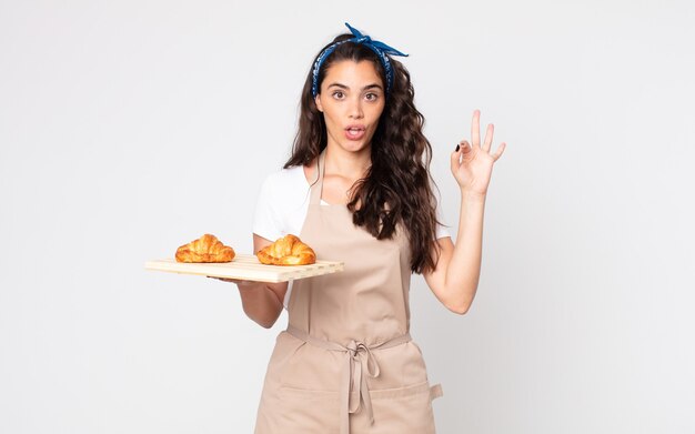 Jolie femme se sentant heureuse, montrant son approbation avec un geste correct et tenant un plateau de croissants