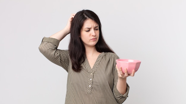 Jolie femme se sentant perplexe et confuse, se grattant la tête tenant un bol de pot vide. agent assistant avec un casque