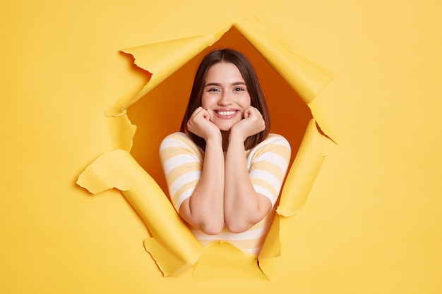 Jolie femme séduisante souriante se tient dans un trou de papier déchiré regardant à travers une percée de fond jaune regarde la caméra en gardant les mains sous le menton