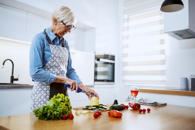 Jolie femme senior blonde en tablier debout dans la cuisine à la maison et couper le poivron jaune