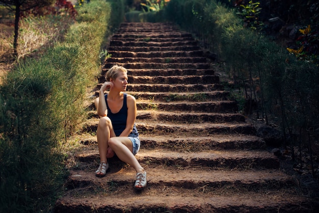 Jolie femme en short en jean, assis sur l'escalier dans le parc d'été.