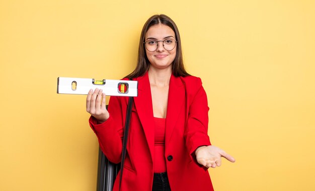 Jolie femme souriant joyeusement amicalement et offrant et montrant un architecte conceptuel avec un niveau