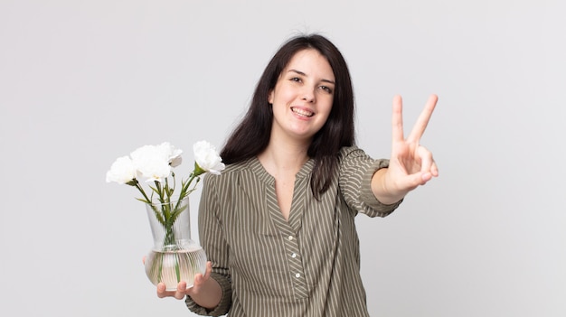 jolie femme souriante et ayant l'air heureuse, gesticulant la victoire ou la paix et tenant des fleurs décoratives. agent assistant avec un casque