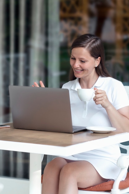 Une jolie femme souriante communique via un ordinateur portable dans un café autour d'une tasse de café dans un café en plein air dans l'après-midi. communication et réseaux sociaux, technologies modernes.