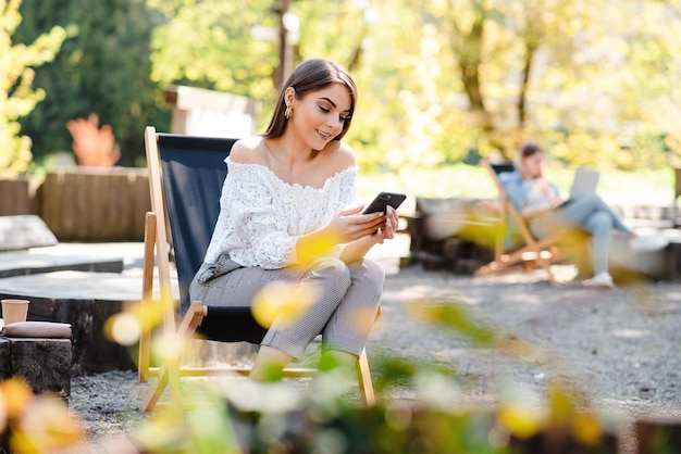 Jolie femme souriante et élégante travaillant à la recherche d'un smartphone indépendant assis dans un parc sur une chaise Centre d'affaires de style urbain de rue, éducation en ligne