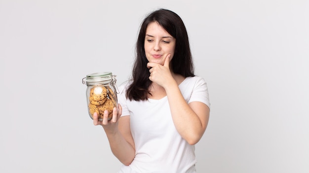 Jolie femme souriante avec une expression heureuse et confiante avec la main sur le menton et tenant une bouteille en verre de cookies