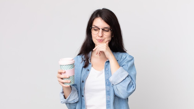 Jolie femme souriante avec une expression heureuse et confiante avec la main sur le menton et tenant un café à emporter