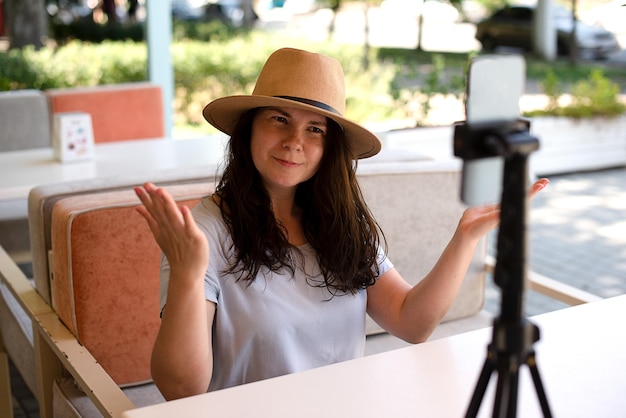 Jolie femme souriante à l'extérieur assise avec un trépied et un smartphone faisant un appel vidéo