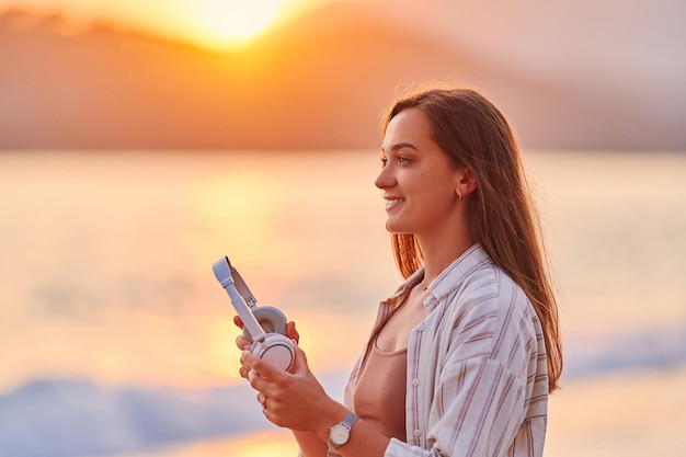 Jolie femme souriante et heureuse aime écouter de la musique de voyage relaxante au bord de la mer au coucher du soleil Beau moment de vie