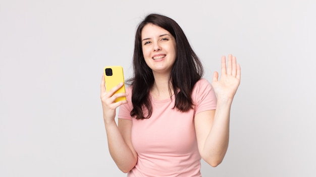 jolie femme souriante joyeusement, agitant la main, vous accueillant et vous saluant à l'aide d'un téléphone intelligent