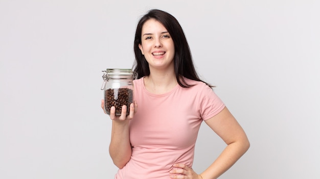 Jolie femme souriante joyeusement avec une main sur la hanche et confiante et tenant une bouteille de grains de café