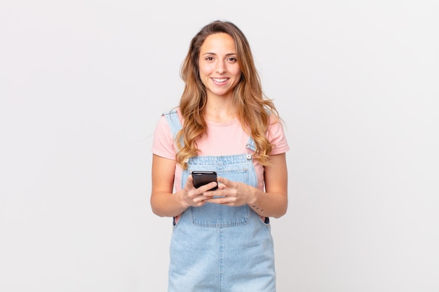 Jolie femme souriante joyeusement avec une main sur la hanche et confiante et tenant un smartphone