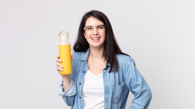 Jolie femme souriante joyeusement avec une main sur la hanche et confiante et tenant un thermos de café