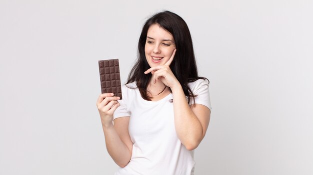 Jolie femme souriante joyeusement et rêvant ou doutant et tenant une barre de chocolat