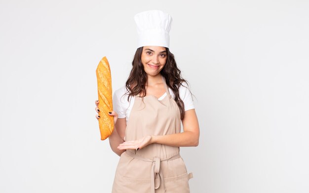 jolie femme souriante joyeusement, se sentant heureuse et montrant un concept portant un tablier et tenant une baguette de pain